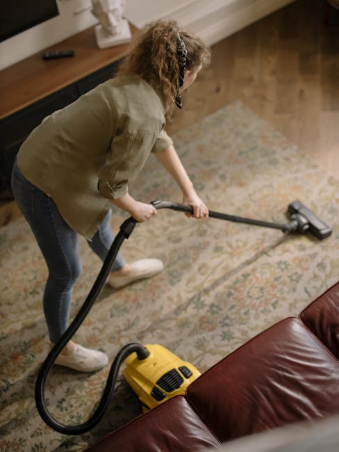 A woman with curly hair, dressed in a beige jacket, blue jeans, and white sneakers, is using a yellow vacuum cleaner with a long hose to perform surface cleaning on a patterned area rug in a living room. She holds the vacuum's hose nozzle directed towards the rug, which is partially situated next to a dark red leather sofa. The room features wooden flooring and a wooden sideboard against the wall with some decor items on top. The space is well-lit with natural light, highlighting the cleanliness and freshness of the rug as part of a professional domestic cleaning process, performed by Highbury Carpet Cleaners, to ensure hygiene and surface maintenance.