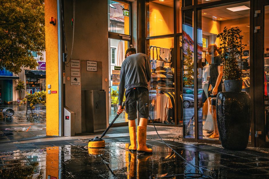 Two maintenance workers dressed in orange and red safety uniforms are engaged in surface cleaning on a busy Highbury street. They are pushing a cleaning cart with equipment, near a pedestrian crossing