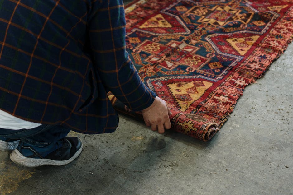 A person wearing a dark blue checkered blazer, white shirt, and sneakers is carefully lifting the corner of a colorful, traditional patterned rug with red, yellow, and blue geometric designs. The rug