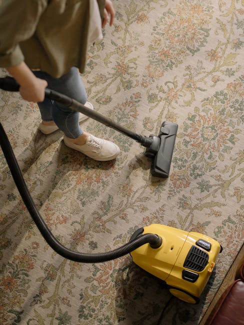 A person wearing a dark blue checkered blazer, white shirt, and sneakers is carefully lifting the corner of a colorful, traditional patterned rug with red, yellow, and blue geometric designs. The rug