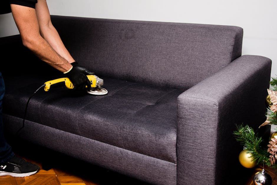 A person wearing black gloves is using an electric handheld upholstery cleaner to deep clean the dark grey fabric sofa in a living room. The sofa features a cushioned backrest and armrest, and the fabric appears slightly damp from the cleaning process. The room has natural lighting, with a portion of a decorated Christmas tree visible on the right side, featuring gold and pink ornaments. The floor is wooden, and the overall scene emphasizes surface cleaning and maintenance typical of residential upholstery sanitisation performed by Highbury Carpet Cleaners for period homes in the Highbury Fields area.