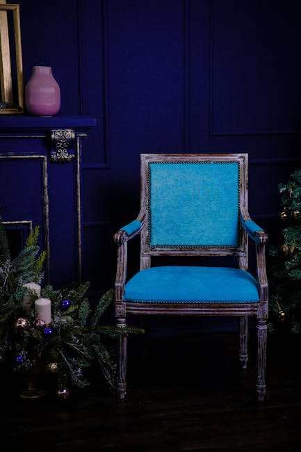 An elegant armchair with a wooden frame and light blue upholstered cushions and arms, positioned against a deep blue paneled wall in a room decorated for the holidays. To the left, a blue mantelpiece holds a pink vase, and around the base of the chair, festive decorations including pine branches, pinecones, and ornaments are visible. The dark wooden floor beneath the chair is clean and polished, reflecting the ambient light. This setting exemplifies a well-maintained, stylish interior where surface cleaning and deep cleaning may have been applied to ensure a tidy and hygienic environment, consistent with the services of Highbury Carpet Cleaners for period homes. The lighting highlights the textures and colors of the furniture and decorations, emphasizing cleanliness and presentation.