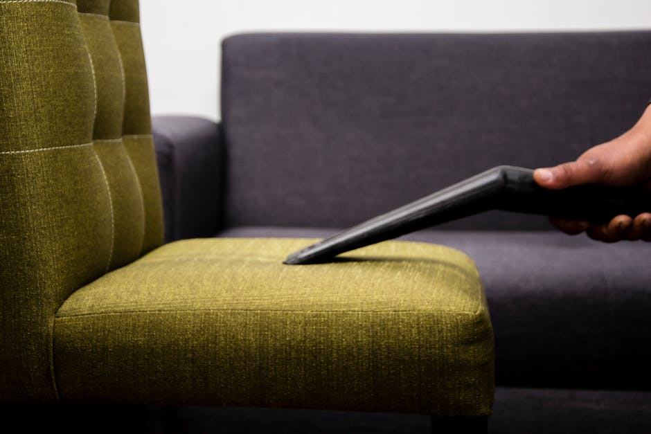 A person wearing black gloves is using an electric handheld upholstery cleaner to deep clean the dark grey fabric sofa in a living room. The sofa features a cushioned backrest and armrest, and the fabric appears slightly damp from the cleaning process. The room has natural lighting, with a portion of a decorated Christmas tree visible on the right side, featuring gold and pink ornaments. The floor is wooden, and the overall scene emphasizes surface cleaning and maintenance typical of residential upholstery sanitisation performed by Highbury Carpet Cleaners for period homes in the Highbury Fields area.