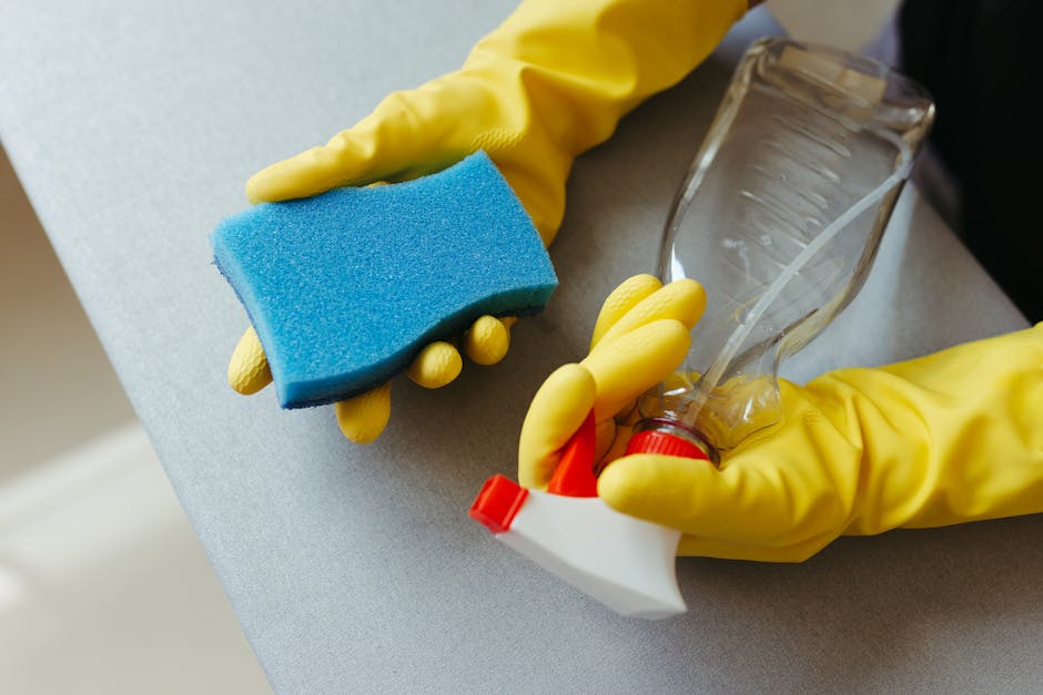 Close-up of a person wearing yellow rubber gloves holding a blue cleaning sponge in one hand and a spray bottle with a transparent plastic covering in the other. The scene shows a smooth, light grey countertop surface in a well-lit room, with parts of the sponge and spray bottle visible. The cleaning supplies are clean and neatly arranged, indicating an active surface cleaning or sanitisation process, typical of domestic or commercial cleaning practices. The background is blurred, emphasizing the tools and the cleanliness of the area, consistent with the professional cleaning services offered by Highbury Carpet Cleaners, especially for tips on avoiding hidden cleaning charges in Highbury.
