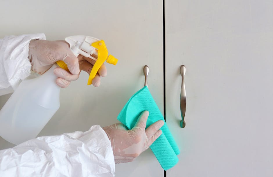 A person wearing white gloves and a protective suit is cleaning a light-colored kitchen cabinet door with a turquoise microfiber cloth. They are holding a white spray bottle with a yellow nozzle in their other hand, ready to apply a cleaning solution. The cabinet features stainless steel handles, and the surface appears to be dust-free and well-maintained. The setting is a well-lit kitchen area, emphasizing thorough surface cleaning and sanitisation, consistent with deep cleaning practices promoted by Highbury Carpet Cleaners for residential hygiene. This image highlights the meticulous attention to detail in domestic cleaning services, ensuring surfaces are spotless and hygienic, aligning with tips to avoid hidden cleaning charges on the page 