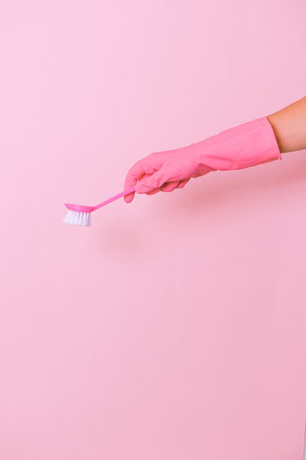 Close-up of a person wearing yellow rubber gloves holding a blue cleaning sponge in one hand and a spray bottle with a transparent plastic covering in the other. The scene shows a smooth, light grey countertop surface in a well-lit room, with parts of the sponge and spray bottle visible. The cleaning supplies are clean and neatly arranged, indicating an active surface cleaning or sanitisation process, typical of domestic or commercial cleaning practices. The background is blurred, emphasizing the tools and the cleanliness of the area, consistent with the professional cleaning services offered by Highbury Carpet Cleaners, especially for tips on avoiding hidden cleaning charges in Highbury.