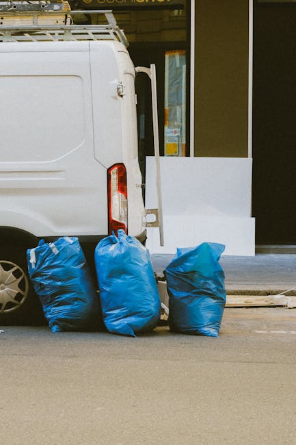 A white commercial van is parked on the street outside a building with large glass windows and a yellow frame, which appears to be a waste disposal or cleaning facility. Next to the van, there are three blue recycling or waste bags placed on the pavement, suggesting waste collection or disposal activity. The window reflection indicates natural daylight, and the scene emphasizes a clean, organized outdoor environment related to waste management. Highbury Carpet Cleaners specializes in surface cleaning and hygiene maintenance, aligning with best practices for waste disposal in compliance with Islington Council regulations for cleaning and waste management. The setting reflects routine waste handling with no visible debris or dirt, highlighting proper sanitation and cleanliness procedures.