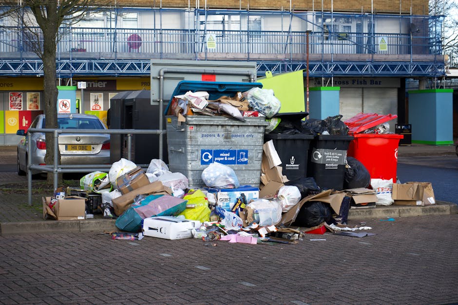 An overflowing public waste disposal area situated on a paved street in front of a commercial building under construction, with a large metal recycling bin and multiple black, red, and green rubbish bags, along with scattered cardboard boxes, paper, and packaging materials on the ground. A silver car is parked behind the waste containers, and street railings separate the area. In the background, the building features scaffolding and a row of shopfronts, including signs indicating a fish bar and other retail outlets. The scene suggests improper waste management and highlights the need for regular cleaning and waste disposal practices in accordance with Islington Council rules, as promoted by Highbury Carpet Cleaners for ensuring proper hygiene and surface cleanliness.