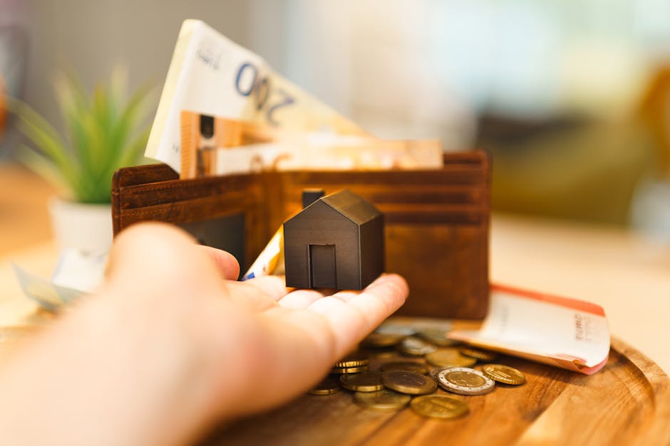 Close-up of a person's hand holding a small black house model over a wooden tray with scattered coins, a brown leather wallet with some paper currency, and a small potted green plant in the background. The scene is well-lit, showcasing a domestic setting related to financial or property matters, consistent with the context of end of tenancy cleaning services. Highbury Carpet Cleaners is mentioned naturally as part of the scene's context for surface cleaning and hygiene maintenance in residential environments.