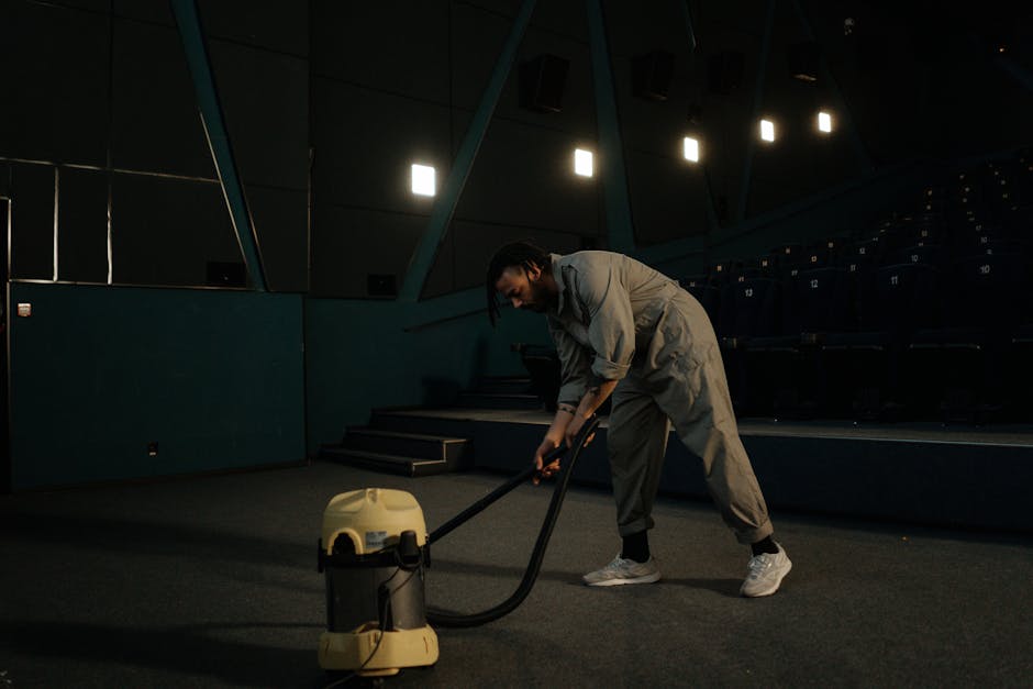 A person operating a yellow industrial carpet cleaner machine in an auditorium or large hall with dark carpeted flooring. The individual is dressed in grey work overalls and white sneakers, leaning forward as they guide the cleaning equipment across the surface. The room features tiered seating with numbered seats visible in the background, and is illuminated by overhead lights, creating a contrast with the darker surroundings. The scene depicts a professional cleaning process, emphasizing surface cleaning and hygiene, consistent with services offered by Highbury Carpet Cleaners for same-day emergency carpet cleaning in Highbury.