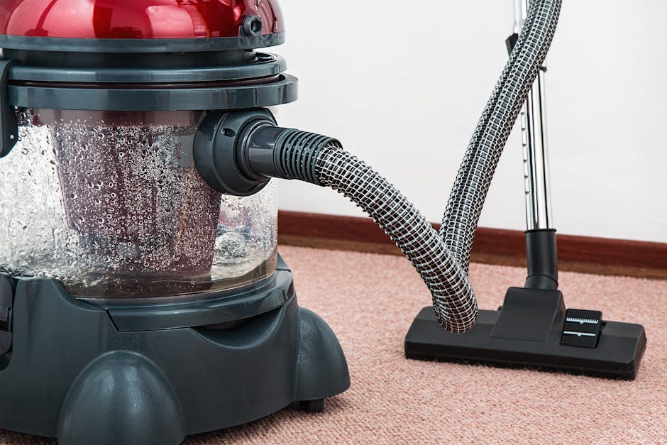 A person operating a yellow industrial carpet cleaner machine in an auditorium or large hall with dark carpeted flooring. The individual is dressed in grey work overalls and white sneakers, leaning forward as they guide the cleaning equipment across the surface. The room features tiered seating with numbered seats visible in the background, and is illuminated by overhead lights, creating a contrast with the darker surroundings. The scene depicts a professional cleaning process, emphasizing surface cleaning and hygiene, consistent with services offered by Highbury Carpet Cleaners for same-day emergency carpet cleaning in Highbury.